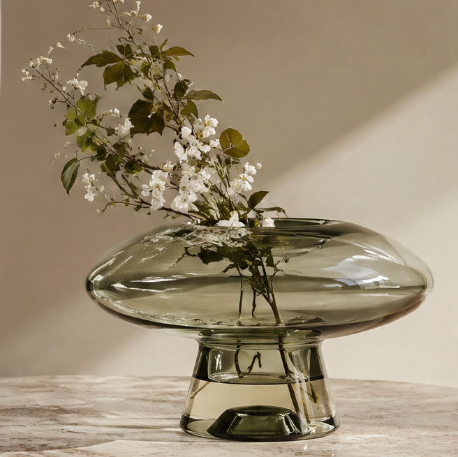 SPIRAVE smoky olive-green saucer-shaped glass vase on a pedestal base, styled with flowering branches